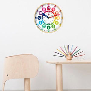 May include: A colourful wooden clock with a rainbow design and numbers around the edge. The clock hands are pointing to 12:00. The clock is hanging on a white wall. There is a wooden table with a wooden peacock pencil holder and a wooden elephant stool in the foreground.