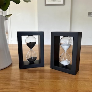 May include: Two black framed hourglass sand timers. One hourglass has black sand and the other has white sand.