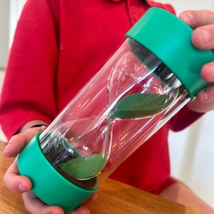 May include: A green hourglass with clear glass bulbs filled with green sand. The hourglass is being held by a person's hand.