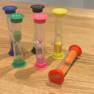 May include: A set of six colourful hourglass timers with different time intervals. The timers are made of glass and have a plastic base. The time intervals are labelled on the base of each timer: 1M, 2M, 3M, 5M, 7M, and 10M.