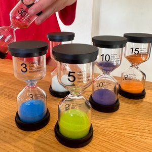 May include: A set of six colourful hourglass sand timers with different time intervals. The timers are made of clear glass with black bases and feature the text "Happy Time Sandglass Minutes" and the corresponding time interval in minutes. The timers are arranged in a row on a wooden surface.