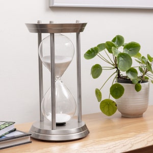 May include: A silver hourglass with white sand, standing on a wooden surface. The hourglass is surrounded by a potted plant with green leaves.