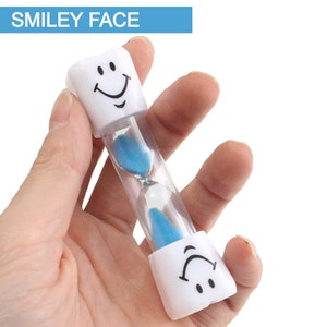 May include: A white hourglass with a smiley face on each end. The hourglass is filled with blue sand and is being held in a hand.