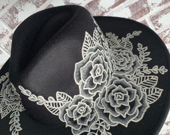 May include: A black wide-brimmed hat featuring intricate white floral embroidery. The design showcases detailed roses and leafy vine patterns that extend from the crown across the brim, creating a high-contrast visual effect on the dark fabric.