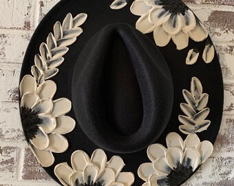 May include: A black wide-brimmed hat featuring a textured, three-dimensional floral design painted in white and cream. The petals and leaves are applied with thick, raised paint strokes, creating a tactile effect around the central crown.