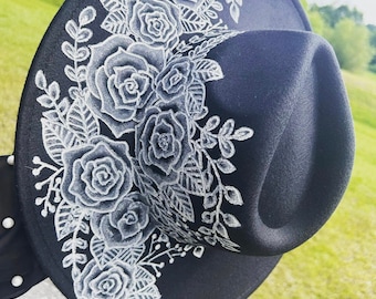 May include: A black wide-brimmed hat featuring a detailed white hand-painted floral design of roses and leaves extending from the brim up to the crown. The hat includes a black band adorned with small white spherical accents on the side.