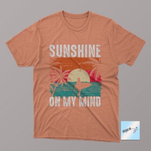 May include: A light orange t-shirt with a vintage-style graphic of a surfer on a wave with palm trees and a sunset. The text "Sunshine on my mind" is printed on the shirt.