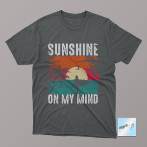 May include: A dark gray t-shirt with a vintage-style graphic of a sunset, palm trees, and a surfer. The text "Sunshine on my mind" is printed in white below the graphic.