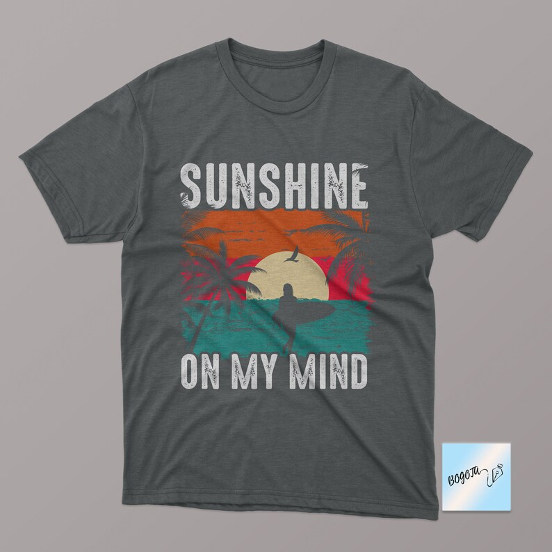 May include: A dark gray t-shirt with a vintage-style graphic of a sunset, palm trees, and a surfer. The text "Sunshine on my mind" is printed in white below the graphic.