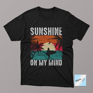 May include: Black t-shirt with a vintage-style graphic of a sunset, palm trees, and a surfer. The text "Sunshine on my mind" is printed below the graphic.
