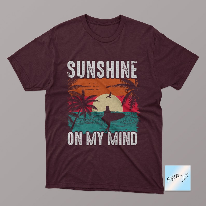 May include: A maroon t-shirt with a vintage-style graphic of a sunset, palm trees, and a surfer. The text "Sunshine on my mind" is printed in white on the shirt.