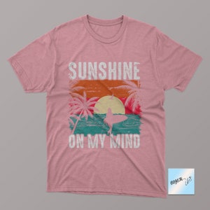 May include: Pink t-shirt with a vintage-style graphic of a sunset, palm trees, and a surfer. The text "Sunshine on my mind" is printed below the graphic.