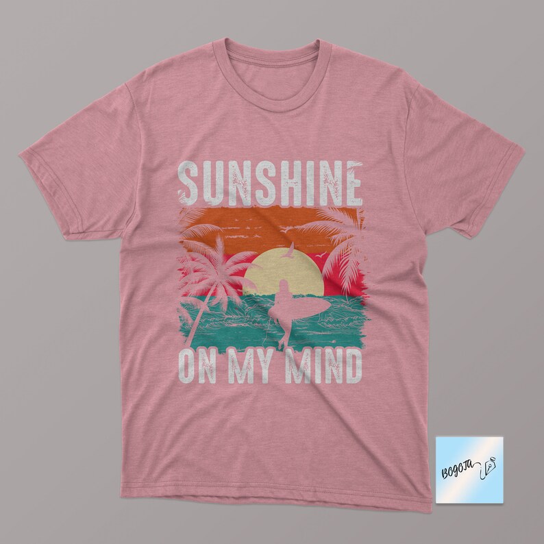May include: Pink t-shirt with a vintage-style graphic of a sunset, palm trees, and a surfer. The text "Sunshine on my mind" is printed below the graphic.