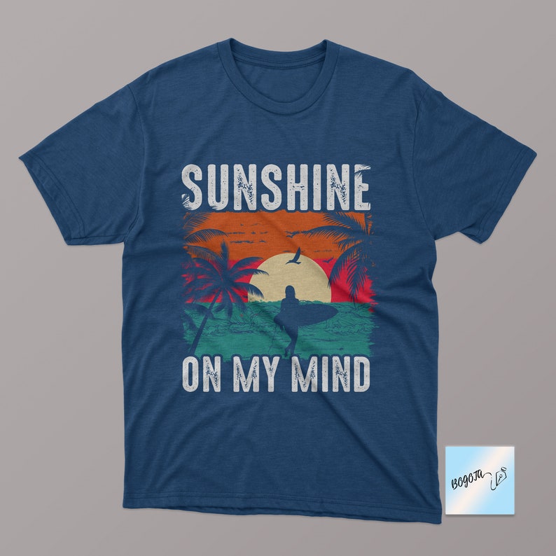 May include: A navy blue t-shirt with a vintage-style graphic of a sunset, palm trees, and a surfer. The text "Sunshine on my mind" is printed in white below the graphic.