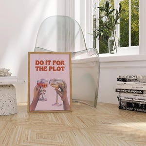 Op de afbeelding: Een ingelijste print met de tekst "DO IT FOR THE PLOT" in oranje. De print toont twee handen die wijnglazen met kleurrijke drankjes vasthouden. De print staat op een houten vloer, in de buurt van een stapel boeken en een doorzichtige stoel.