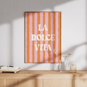 Op de afbeelding: Een ingelijste kunstdruk met een verticaal streepjespatroon in oranje en roze. De woorden "LA DOLCE VITA" staan in het wit, gecentreerd op de print. De print wordt weergegeven op een houten oppervlak met een klein boek en een vaas.