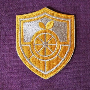 May include: Embroidered shield-shaped patch with a yellow and silver design. The design features a stylized orange with leaves and a wheel. The patch is set against a purple fabric background.