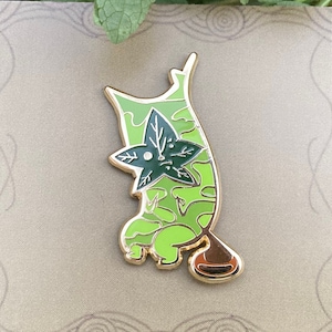 May include: A green and gold enamel pin featuring a cartoon creature with a leafy design. The creature has a large, round head with a single eye and a small, pointed mouth. The pin is attached to a brown backing card.