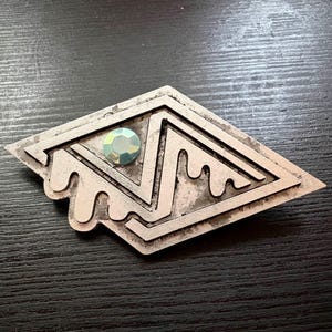 Peut inclure: Broche en métal argenté avec un motif géométrique et une pierre précieuse irisée au centre. La broche a une forme de losange avec des détails texturés et superposés et un bord stylisé et fluide. La pierre précieuse ajoute une touche de couleur et d'éclat.