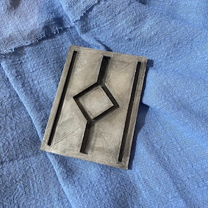 Könnte beinhalten: Ein graues, rechteckiges, 3D-gedrucktes Objekt mit einem geometrischen Design. Das Objekt hat eine erhabene Diamantform in der Mitte mit Linien, die sich vom Diamanten zu den Rändern des Objekts erstrecken.