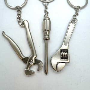 Puede incluir: Tres llaveros plateados con diseños de herramientas. Los llaveros presentan una llave inglesa, un destornillador y un alicate.