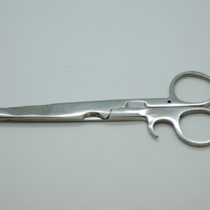Könnte beinhalten: Eine chirurgische Schere aus Edelstahl mit einer langen, dünnen Klinge und einem Ringgriff. Die Schere ist geschlossen und liegt auf einer weißen Oberfläche.