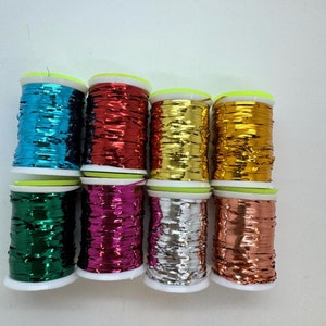 Op de afbeelding: Acht spoelen metallic lint in verschillende kleuren, waaronder blauwgroen, rood, goud, brons, groen, magenta, zilver en een donkerder goud. Elke spoel heeft een witte basis en een neon groene bovenkant.