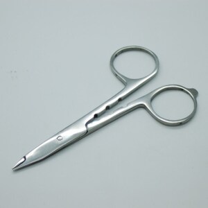 Könnte beinhalten: Eine kleine, silberne Schere mit einer spitzen Spitze und einer gezahnten Klinge. Die Schere ist geschlossen und liegt auf einer weißen Oberfläche.