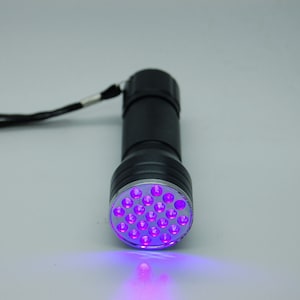 Könnte beinhalten: Eine schwarze Taschenlampe mit einem violetten LED-Licht. Die Taschenlampe hat einen schwarzen Gurt und befindet sich auf einer weißen Oberfläche.