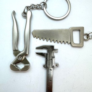 Peut inclure: Un porte-clés argenté avec une scie, une pince et un pied à coulisse. Le porte-clés a un anneau et une chaîne argentés.