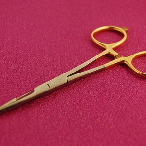 Könnte beinhalten: Eine goldfarbene chirurgische Pinzette mit einem silberfarbenen Griff. Die Pinzette ist geschlossen und liegt auf einer rosa Oberfläche.