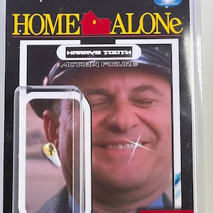 May include: A plastic action figure packaging for a Home Alone Harry's Tooth action figure. The packaging features a close-up image of the character, Harry, with a gold tooth. The text on the packaging reads "Harry's Tooth Action Figure", "Limited Hand Made Gold Tooth", and "Excuse me are you parents home?"