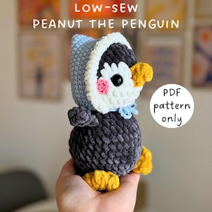 May include: A handmade crochet penguin named Peanut, with a blue and white hood, pink cheeks, and yellow feet. The penguin is held in a hand. Text on the image reads "LOW-SEW PEANUT THE PENGUIN" and "PDF pattern only."
