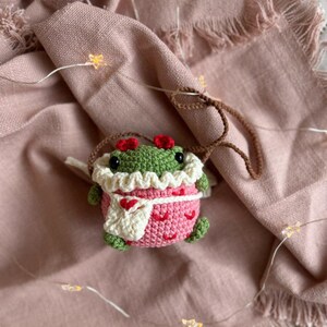 May include: A crocheted frog wearing a pink dress with white trim and a white crocheted scarf. The frog is sitting on a pink fabric background with white lights.