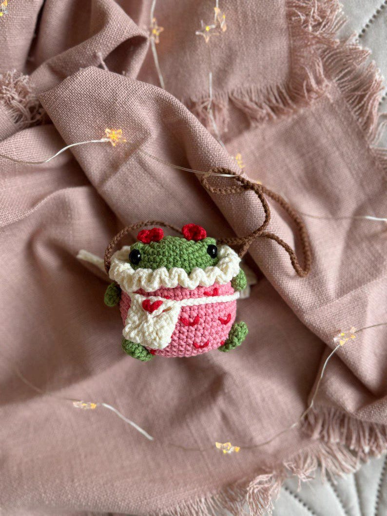 May include: A crocheted frog wearing a pink dress with white trim and a white crocheted scarf. The frog is sitting on a pink fabric background with white lights.