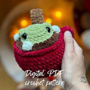 May include: A crochet pattern for a plush toy in the shape of a cup of hot chocolate with a marshmallow on top. The cup is red, the hot chocolate is brown, and the marshmallow is white. The toy is being held by a hand wearing a silver bracelet. The text "Digital PDF crochet pattern" is visible in the image.