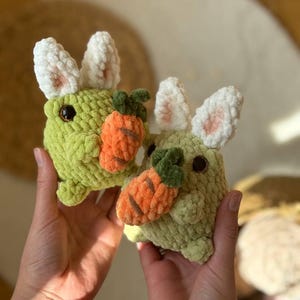 Pu&ograve; includere: Due peluche di coniglio di uncinetto verde che tengono peluche di carota arancione. I conigli hanno orecchie di uncinetto bianche.
