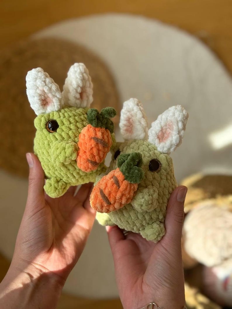 Pu&ograve; includere: Due peluche di coniglio di uncinetto verde che tengono peluche di carota arancione. I conigli hanno orecchie di uncinetto bianche.
