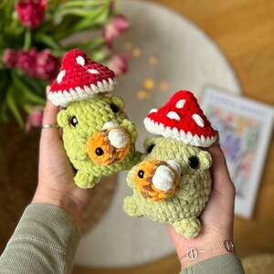 Puede incluir: Dos peluches de hongo verde de crochet con gorros de hongo rojo y blanco. Los peluches tienen ojos naranja y blanco y una nariz blanca.