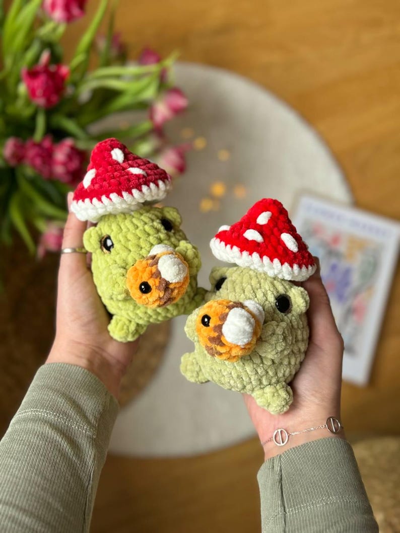 Puede incluir: Dos peluches de hongo verde de crochet con gorros de hongo rojo y blanco. Los peluches tienen ojos naranja y blanco y una nariz blanca.