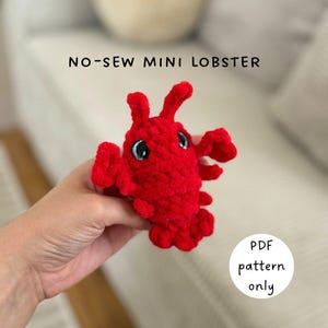 May include: A small, red, crocheted lobster with blue eyes. The text "NO-SEW MINI LOBSTER" is above the lobster, and a white circle with "PDF pattern only" is on the right. The lobster is held in a hand.