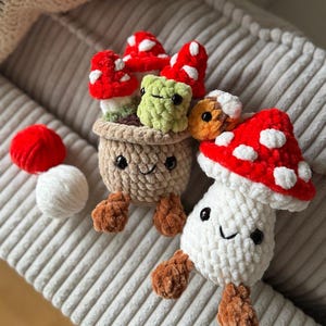 May include: Crocheted mushroom plushies in red, white, and brown. One mushroom is in a pot with a smiling face, and others are scattered around. The mushrooms have white spots on their red caps. The plushies are on a gray corduroy surface.