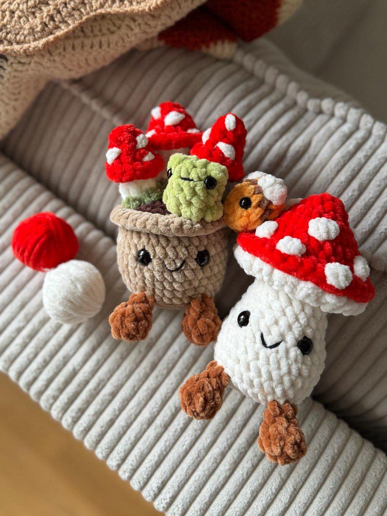 May include: Crocheted mushroom plushies in red, white, and brown. One mushroom is in a pot with a smiling face, and others are scattered around. The mushrooms have white spots on their red caps. The plushies are on a gray corduroy surface.