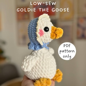 May include: A handmade, white crochet goose toy named "Goldie the Goose" wearing a blue bonnet with a white trim. The goose has a yellow beak and feet, and pink cheeks. The text "LOW-SEW GOLDIE THE GOOSE" and "PDF pattern only" are visible.