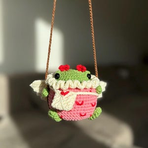 May include: A crocheted frog wearing a pink dress with white hearts and a white scarf. The frog has red flowers on its head and is hanging from a brown string.