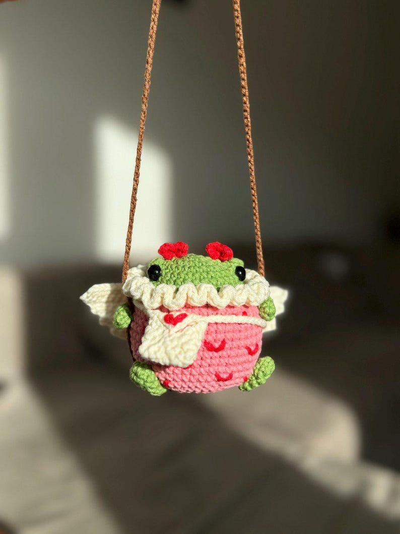 May include: A crocheted frog wearing a pink dress with white hearts and a white scarf. The frog has red flowers on its head and is hanging from a brown string.
