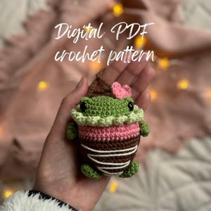 May include: A crocheted frog amigurumi toy with a pink bow, green body, brown and white striped bottom, and pink and white accents. The toy is being held in a hand. Digital PDF crochet pattern.