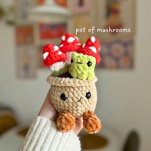 May include: A crocheted pot of mushrooms with a smiling face and orange feet. The pot is beige, and the mushrooms are red with white spots. The text "pot of mushrooms" is in the image.