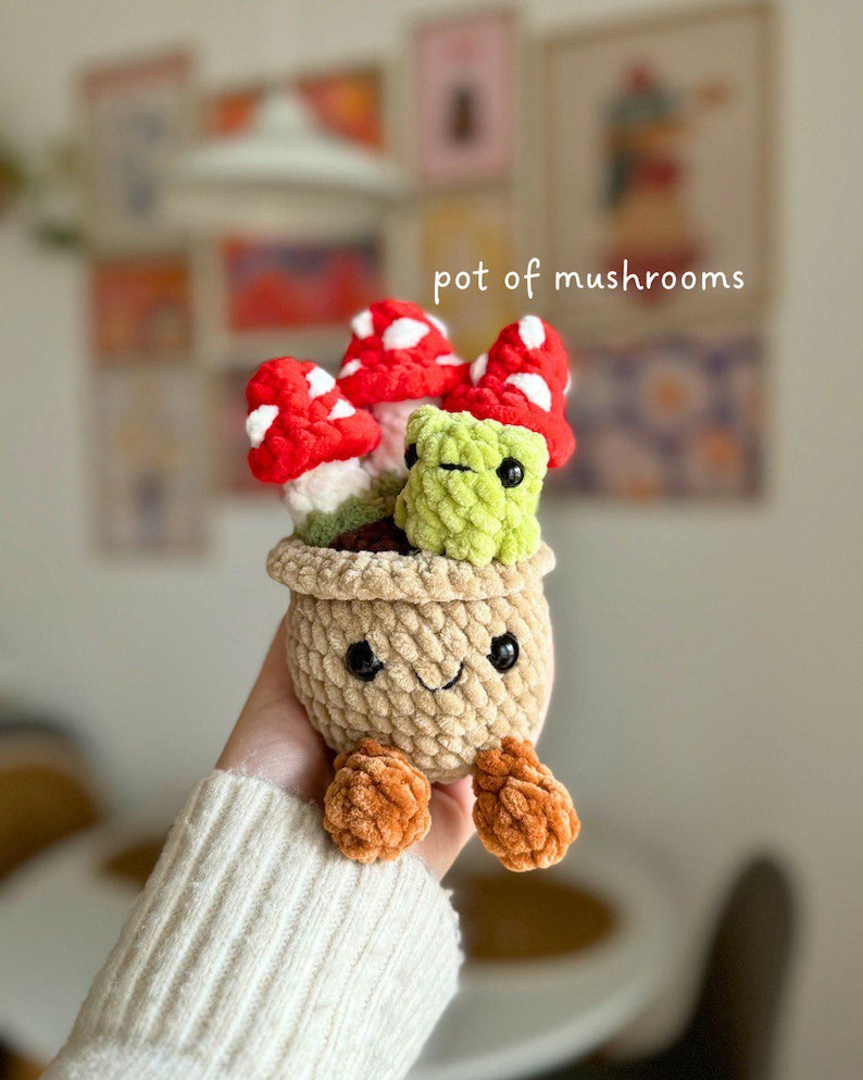May include: A crocheted pot of mushrooms with a smiling face and orange feet. The pot is beige, and the mushrooms are red with white spots. The text "pot of mushrooms" is in the image.