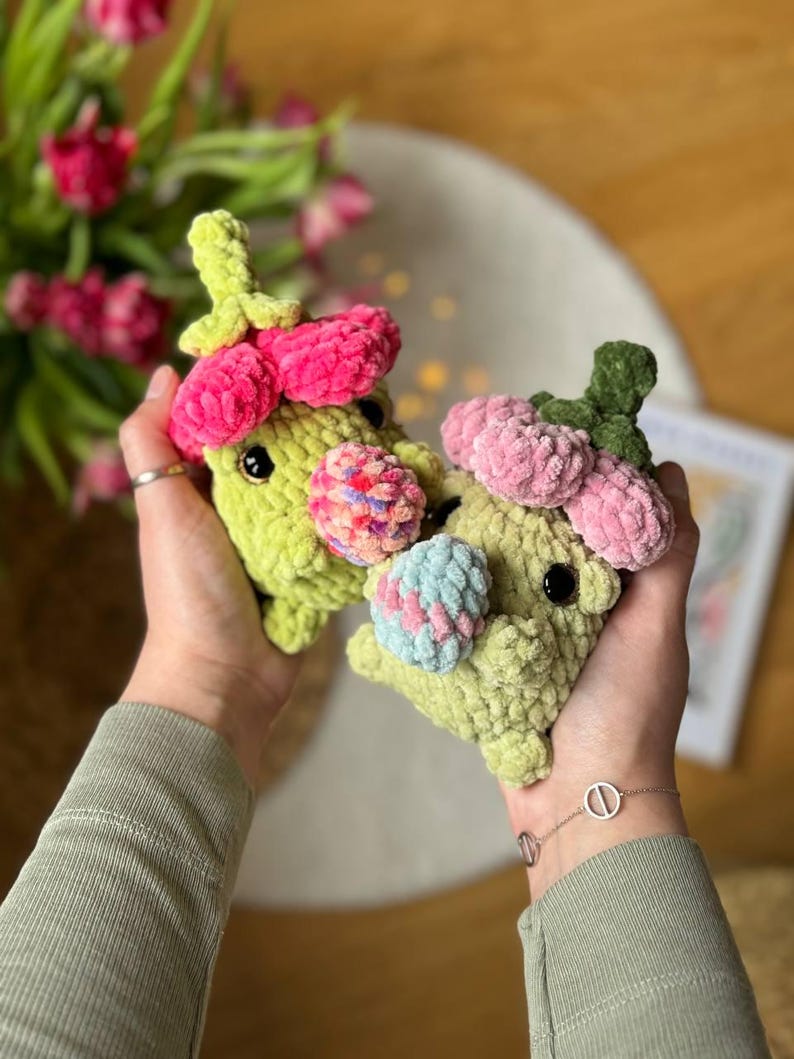 May include: Two crocheted plush toys shaped like flowers with green bodies and pink petals. The toys are holding colorful crocheted balls.